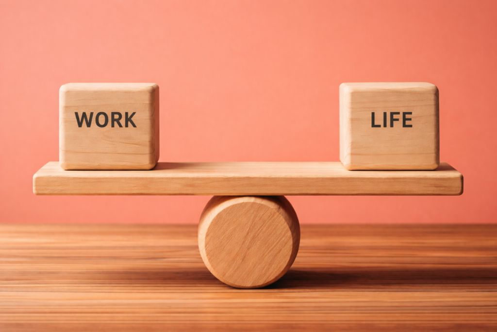 Work life balance concept image showing wooden scales balancing work and life on a coral background, representing wellbeing, boundaries, and sustainable performance.